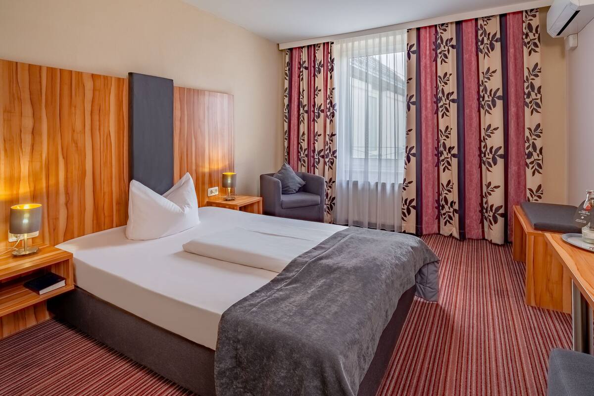A hotel room with a bed, a desk, a chair, a nightstand, and a window with curtains.