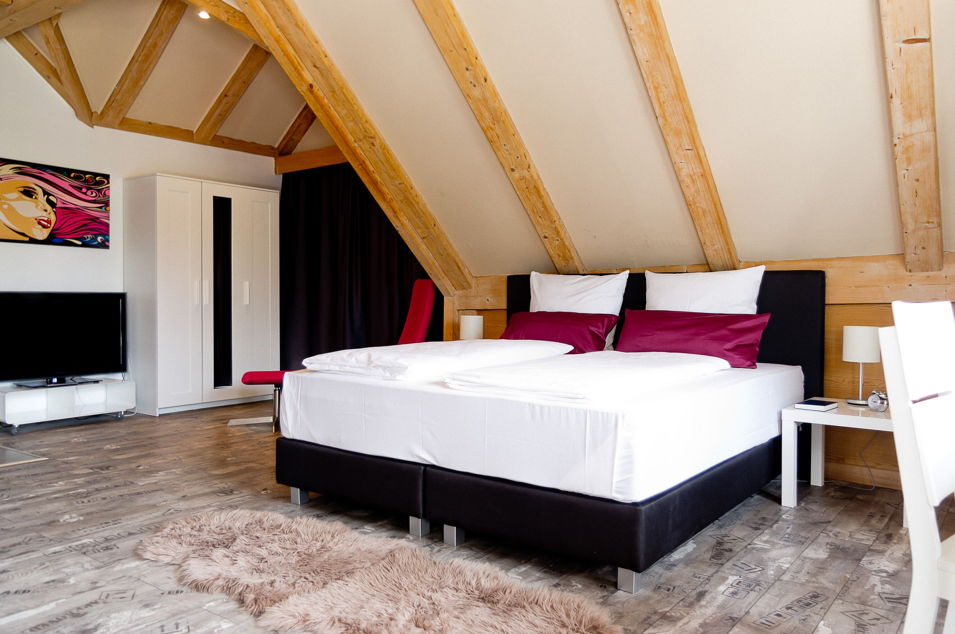 A bedroom with a large bed, wooden ceiling beams, a TV, and a fluffy rug.