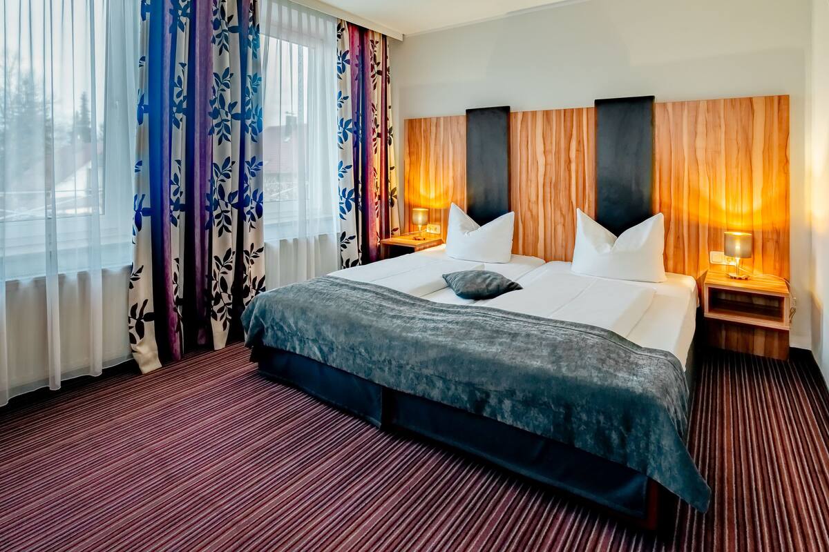 A hotel room with a large bed, two bedside tables with lamps, a headboard with vertical wood panels, and a view of the outdoors through sheer curtains.