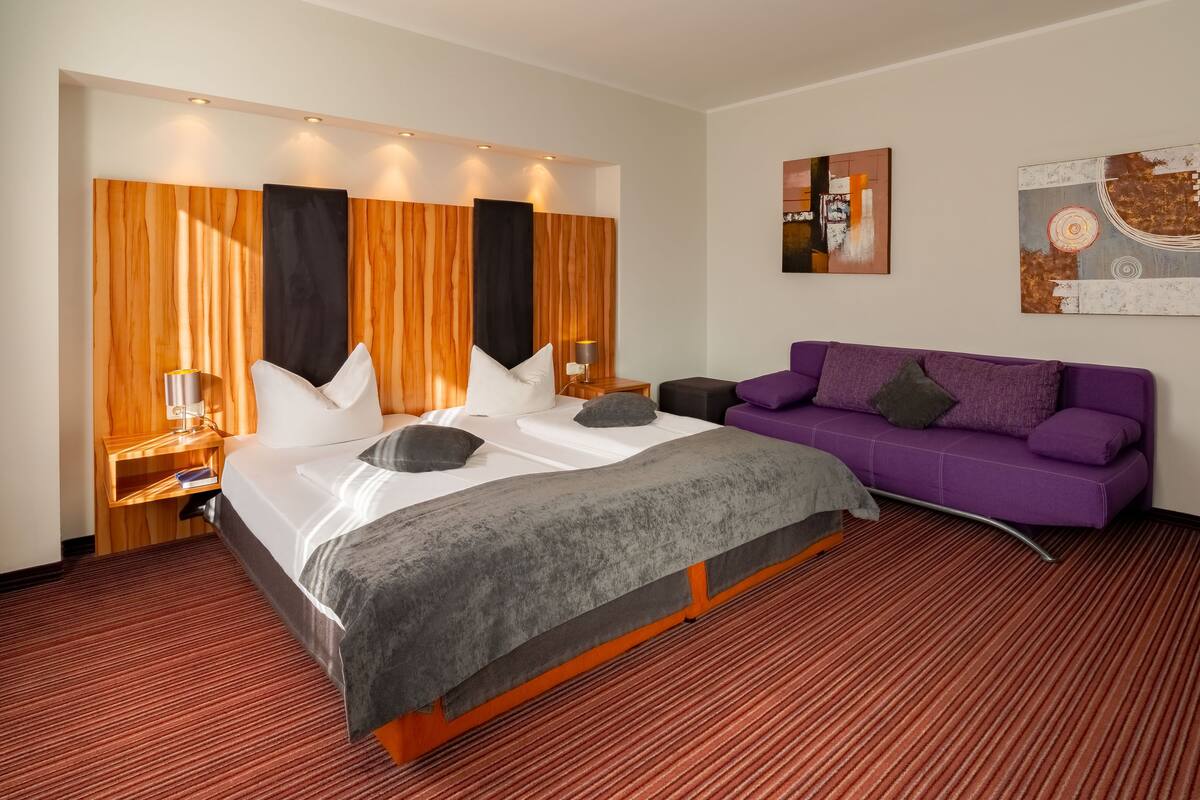 A hotel room with a large bed, a purple sofa, wooden panel walls, and artwork on the walls.