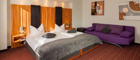 A hotel room with a large bed, a purple sofa, wooden panel walls, and artwork on the walls.