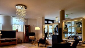 A modern living room with a flat-screen TV, a wooden cabinet, a black armchair, a glass display case with decorative items, and a large chandelier. - Arthotel ANA Style (Augsburg)