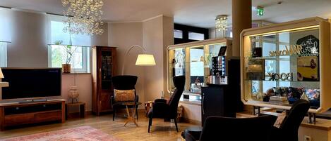 A modern living room with a flat-screen TV, a wooden cabinet, a black armchair, a glass display case with decorative items, and a large chandelier.