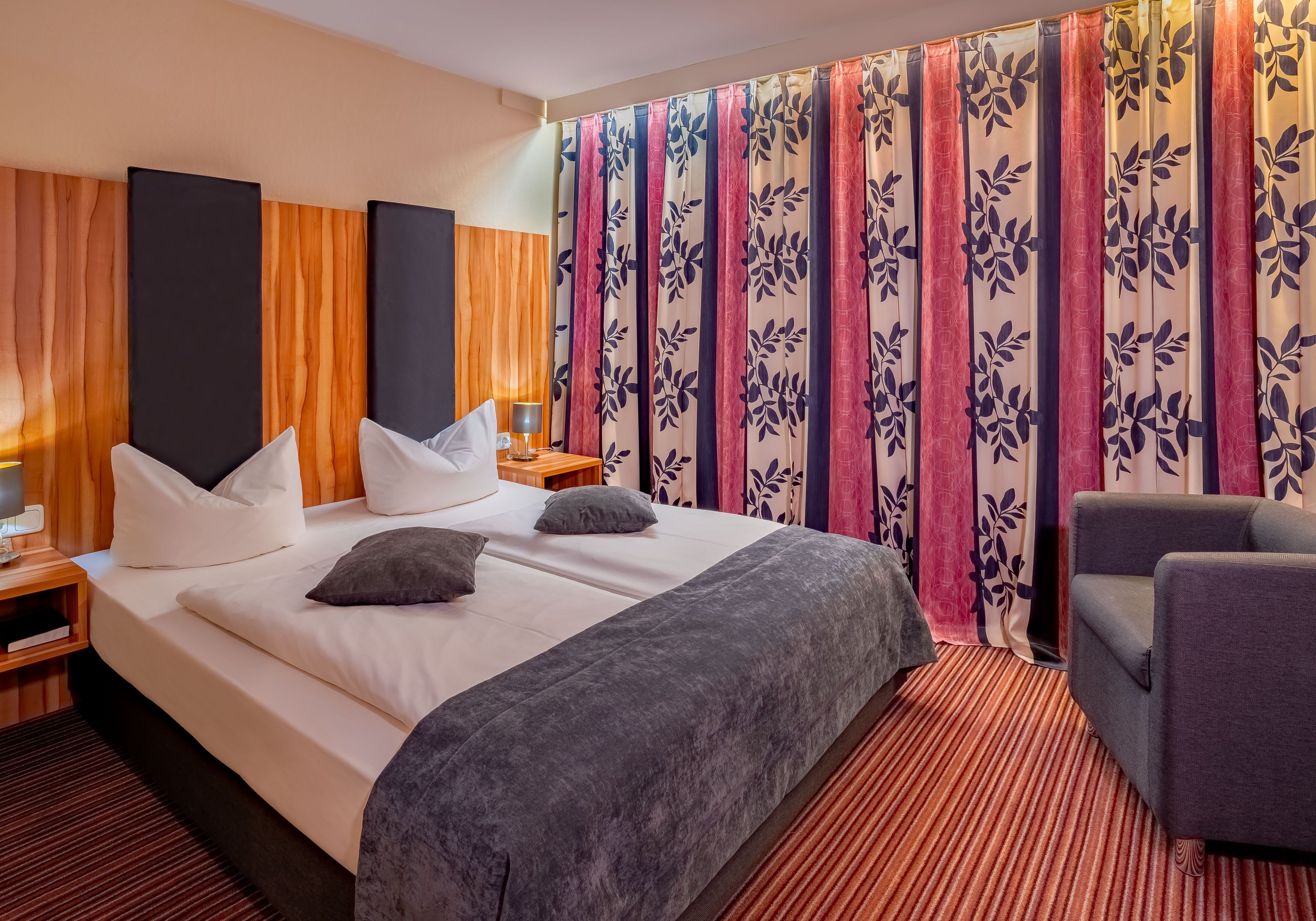Een hotelkamer met een groot bed, twee nachtkastjes, een stoel en een gordijn met een bloemenprint.