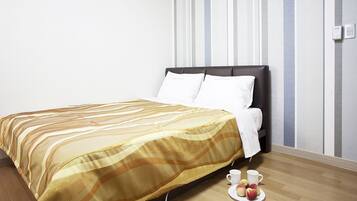 Una cama bien hecha con una colcha a rayas, un cabecero oscuro y almohadas blancas. Hay un plato con manzanas y dos tazas blancas en el suelo, junto a la cama.