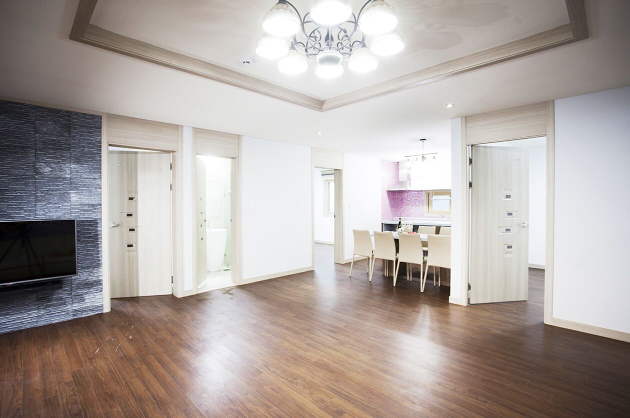 A spacious living area with a flat-screen TV mounted on a textured wall, a dining area with a table and chairs, and a kitchen visible through an open door.