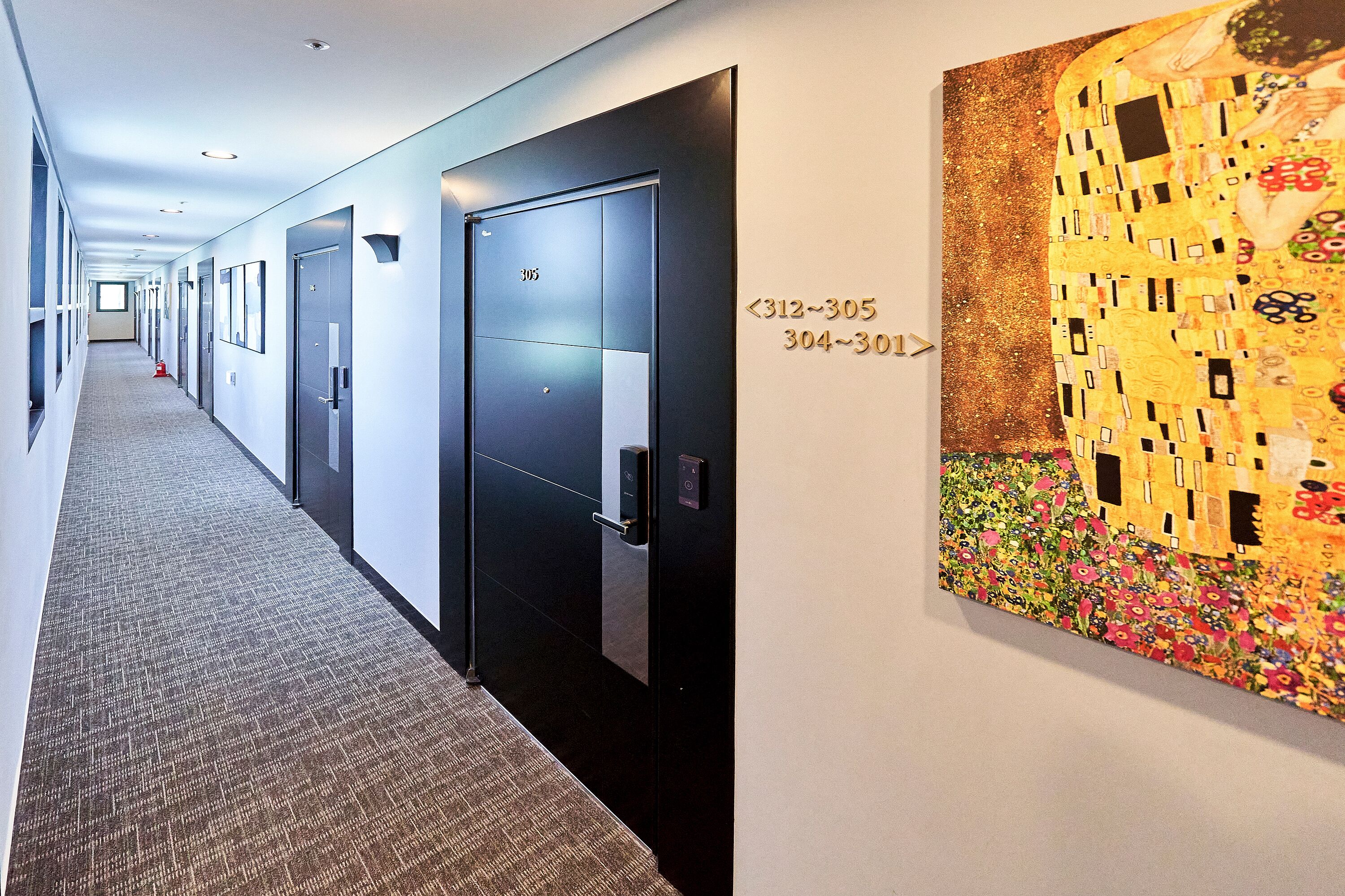 A hotel corridor with doors numbered 312-305 and 304-301, a modern elevator, and a large Gustav Klimt painting on the wall.