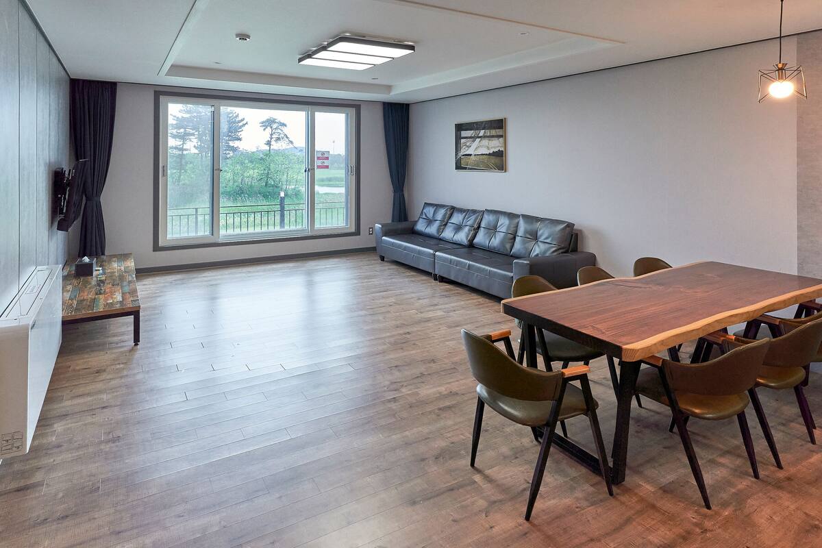 Una habitación amplia con una mesa de comedor de madera, seis sillas, un sofá gris y un ventanal con vistas a zonas verdes.