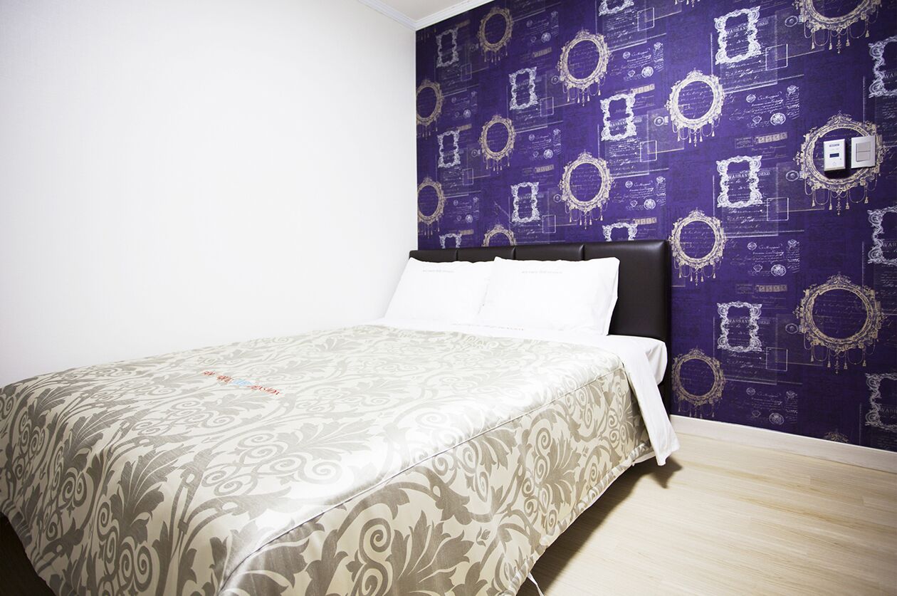 A neatly made bed with a patterned bedspread, a dark headboard, and a purple wallpaper with ornate designs.