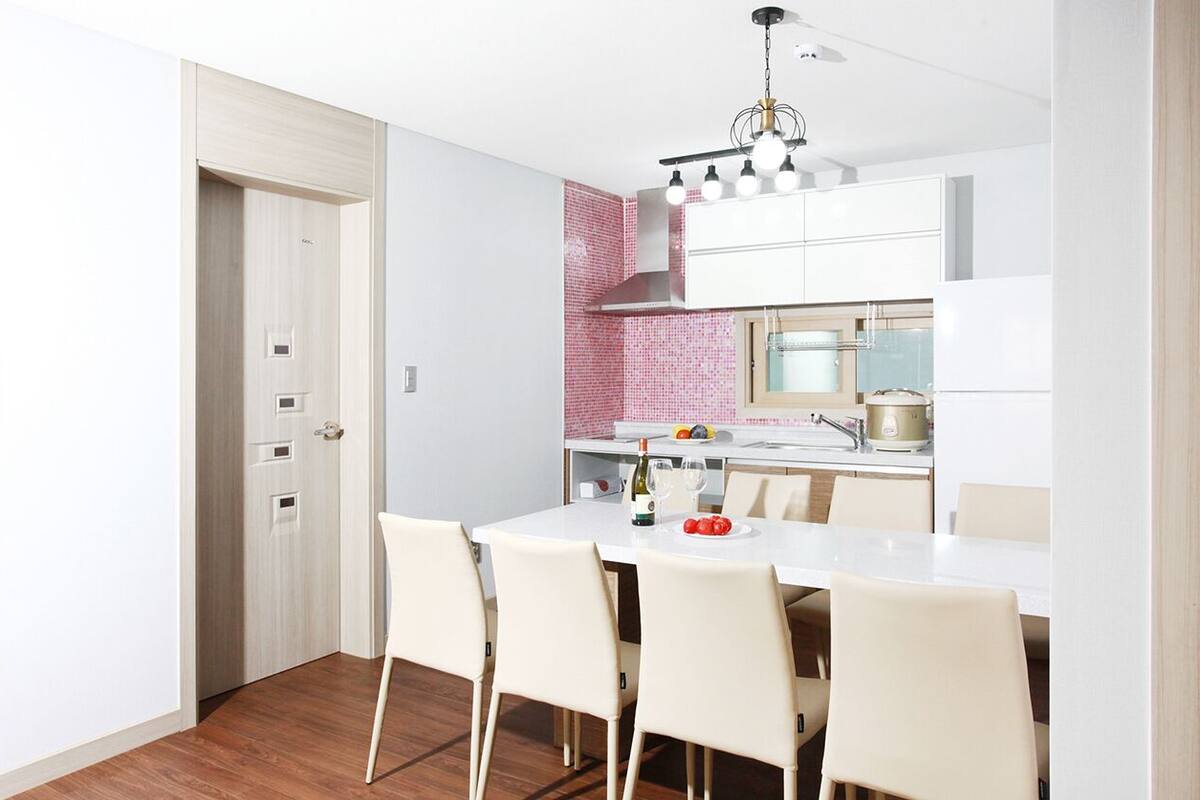 Una cocina moderna con armarios blancos, un salpicadero rosa y una zona de comedor con mesa y sillas blancas.