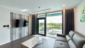 Ein modernes Wohnzimmer mit einem großen Flachbildfernseher an einer Wand aus Holzpaneelen, einem dunkelbraunen Ledersofa, einem Marmortisch und einer großen Schiebeglastür, die auf einen Balkon mit Blick auf Grünflächen und Gebäude führt.