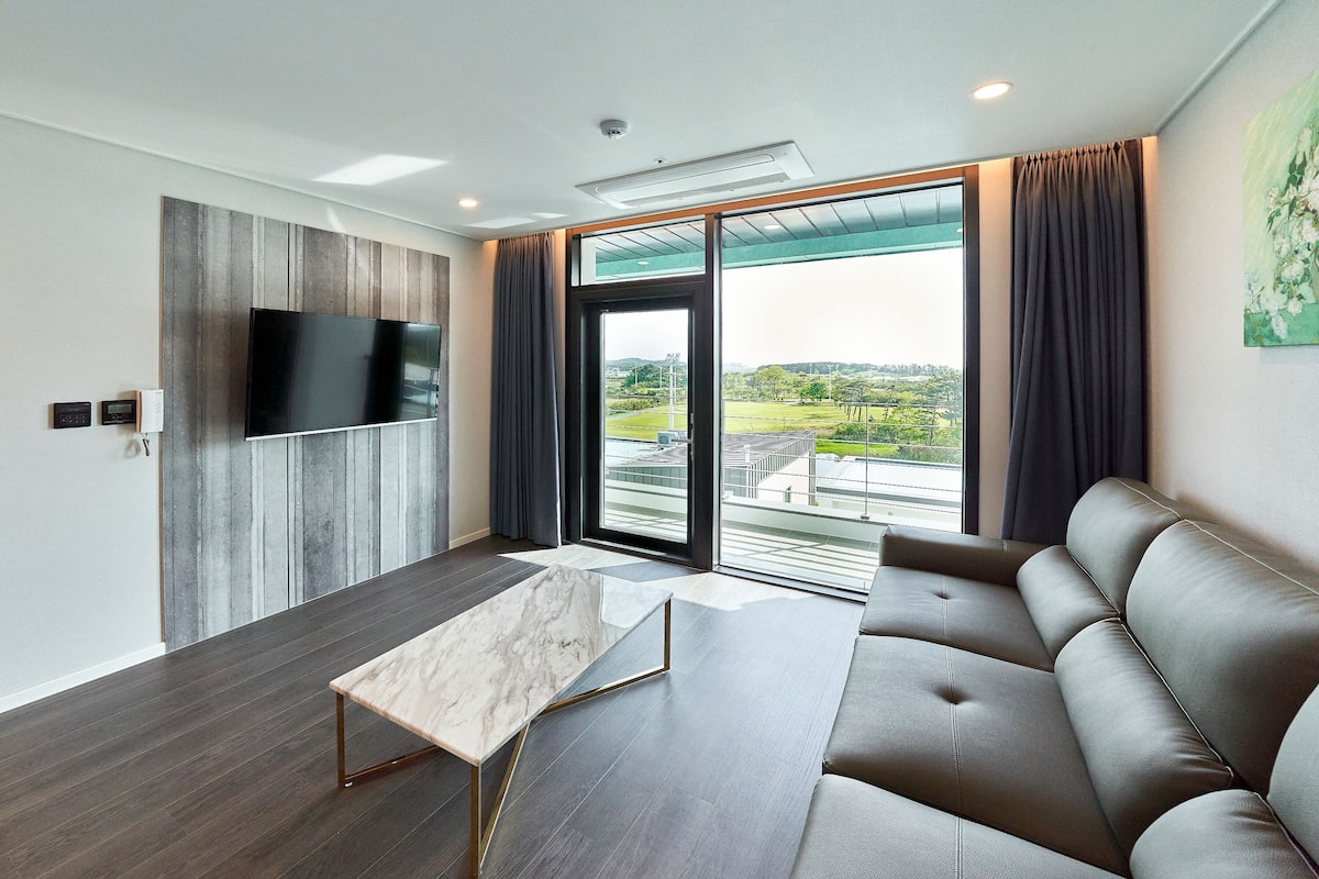 Un moderno salón con una amplia televisión de pantalla plana montada en una pared de paneles de madera, un sofá de cuero oscuro, una mesa de centro de mármol y un gran ventanal corredero que da a un balcón exterior con vistas a zonas verdes y edificios.