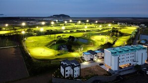 Ein Golfplatz im Abenddämmerung mit beleuchteten Lichtern, einem Teich und modernen Gebäuden inklusive Parkplatz.