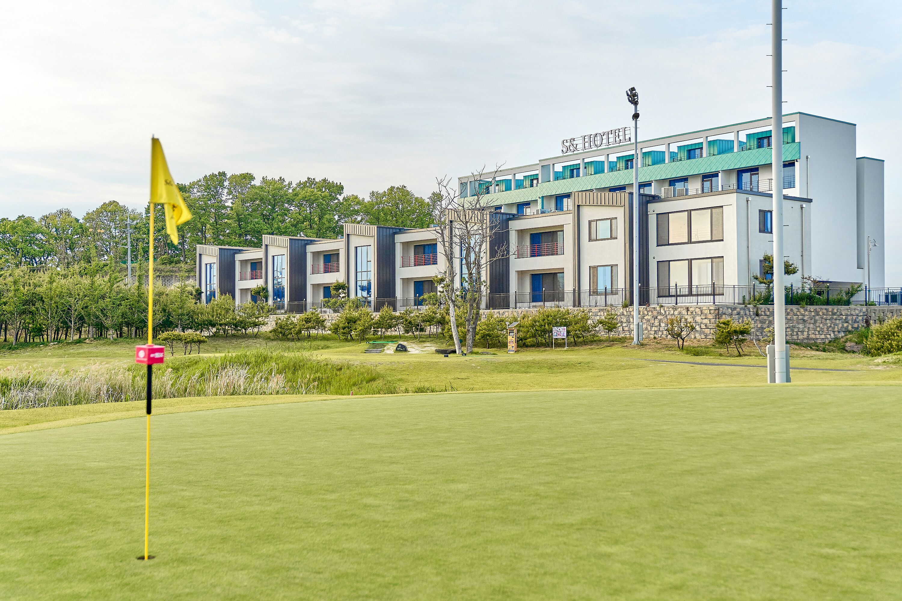 A modern hotel with a flat roof and multiple balconies, surrounded by well-maintained lawns and trees.