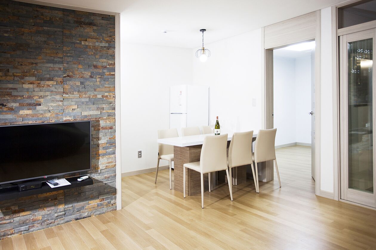 A modern living room with a flat-screen TV on a stone wall, a dining area with a white table and chairs, and a glass door leading to another room.