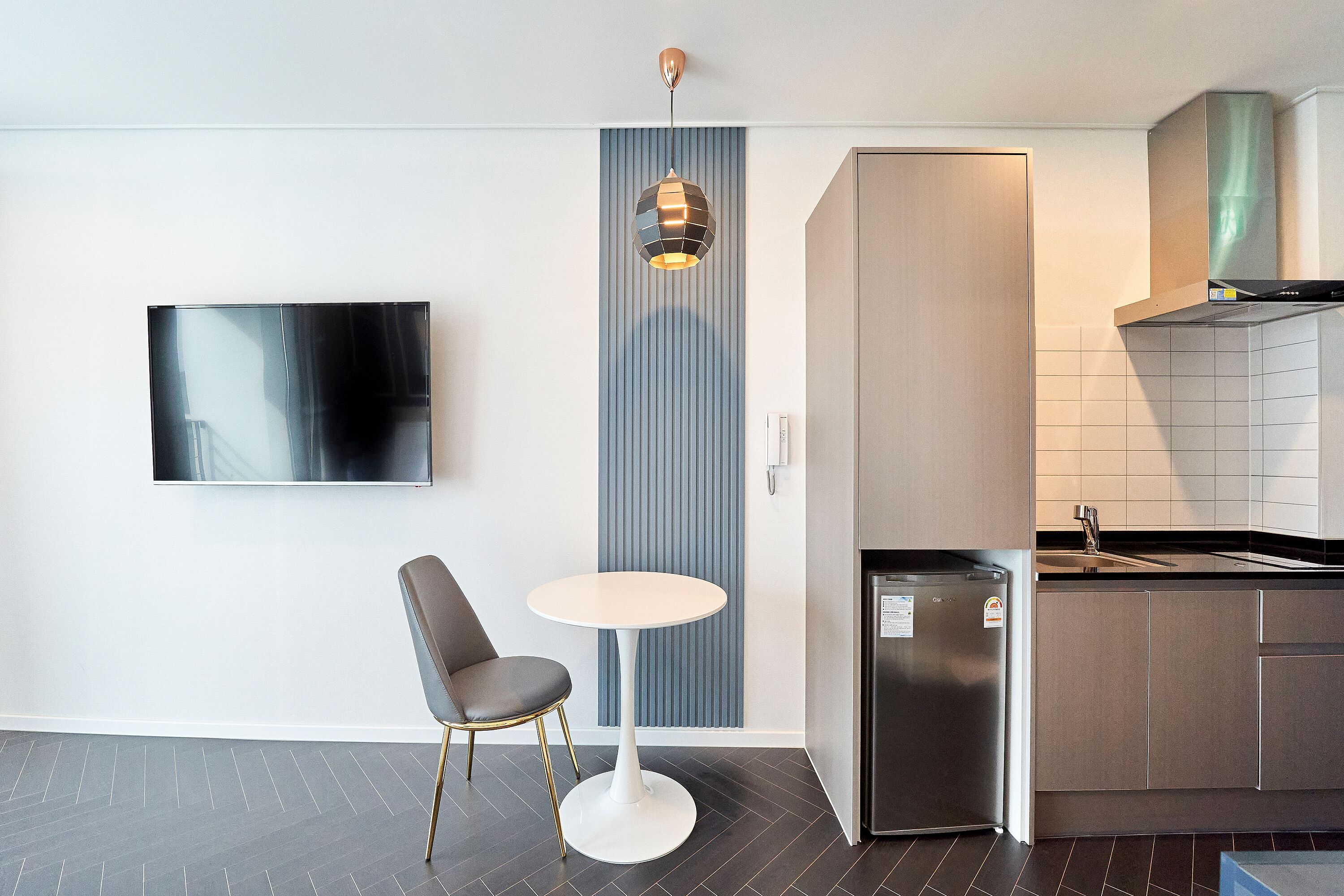 A modern kitchen with a built-in refrigerator, a dining area with a round table and a chair, and a wall-mounted TV.