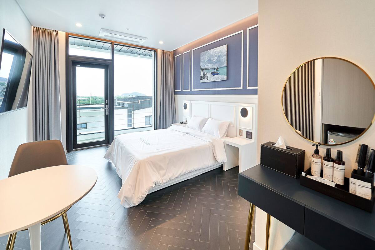 Habitación de hotel con cama, espejo redondo, mesita de noche con artículos de tocador, ventana con cortinas y vistas al exterior.