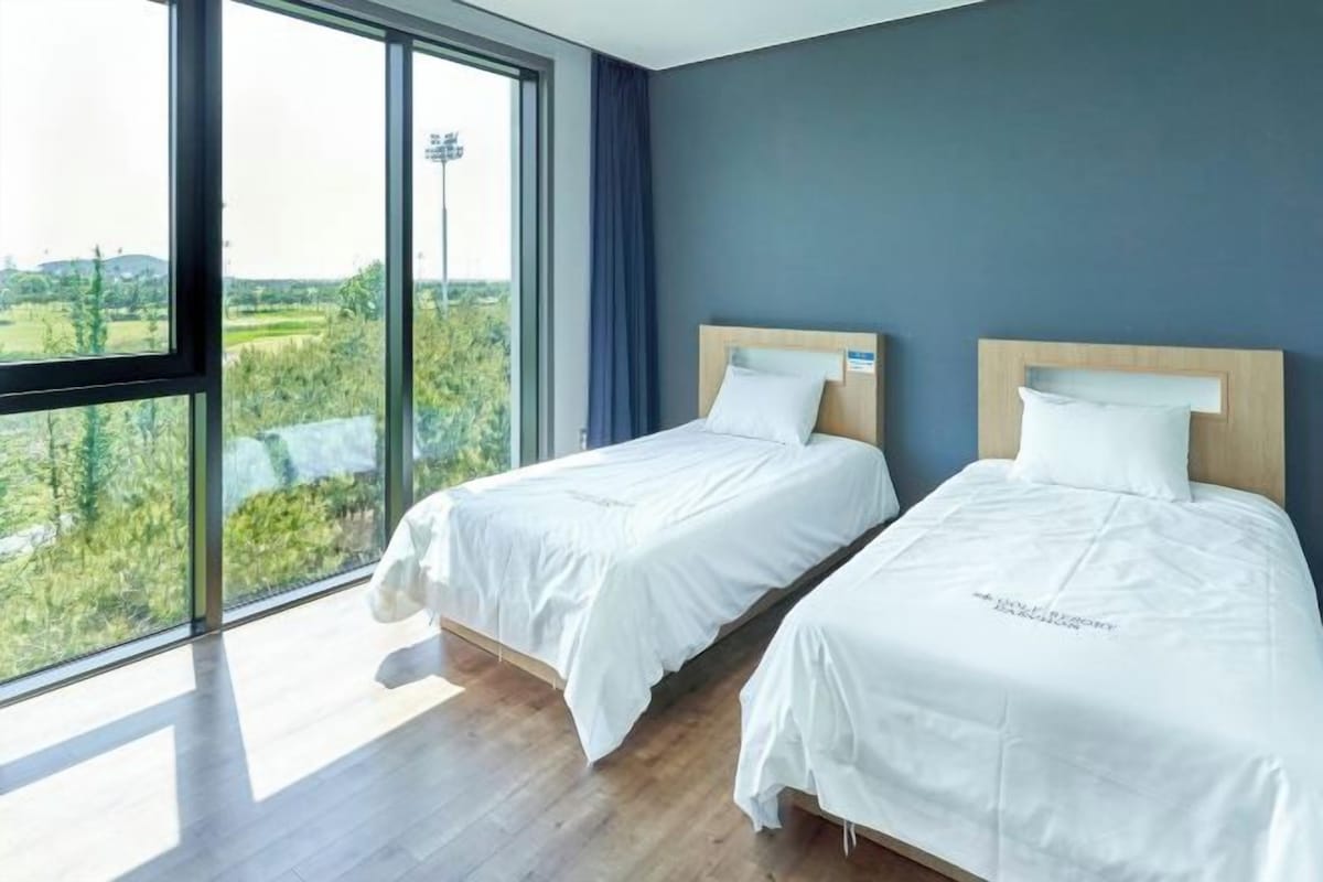 A hotel room with two beds, large windows, and a view of greenery.