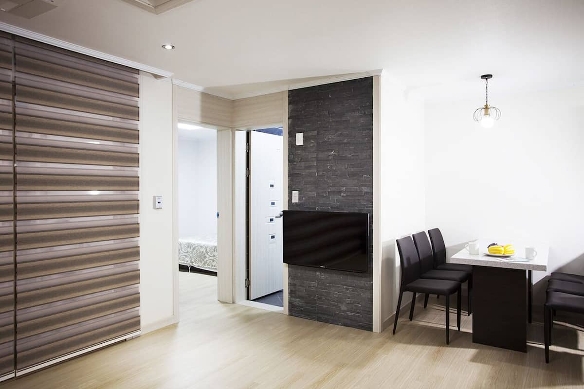 Una habitación de hotel moderna con una puerta corredera, un televisor de pantalla plana montado en una pared oscura, una zona de comedor con una mesa y sillas, y una lámpara en el techo.