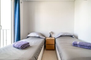 Une petite chambre propre, avec deux lits simples, une table de chevet avec une lampe et une fenêtre avec des rideaux.