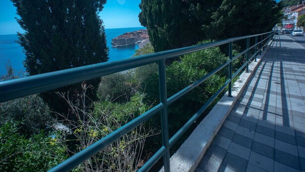 Un paseo marítimo con barandilla de metal, con vista a una masa de agua y una ciudad con edificios.