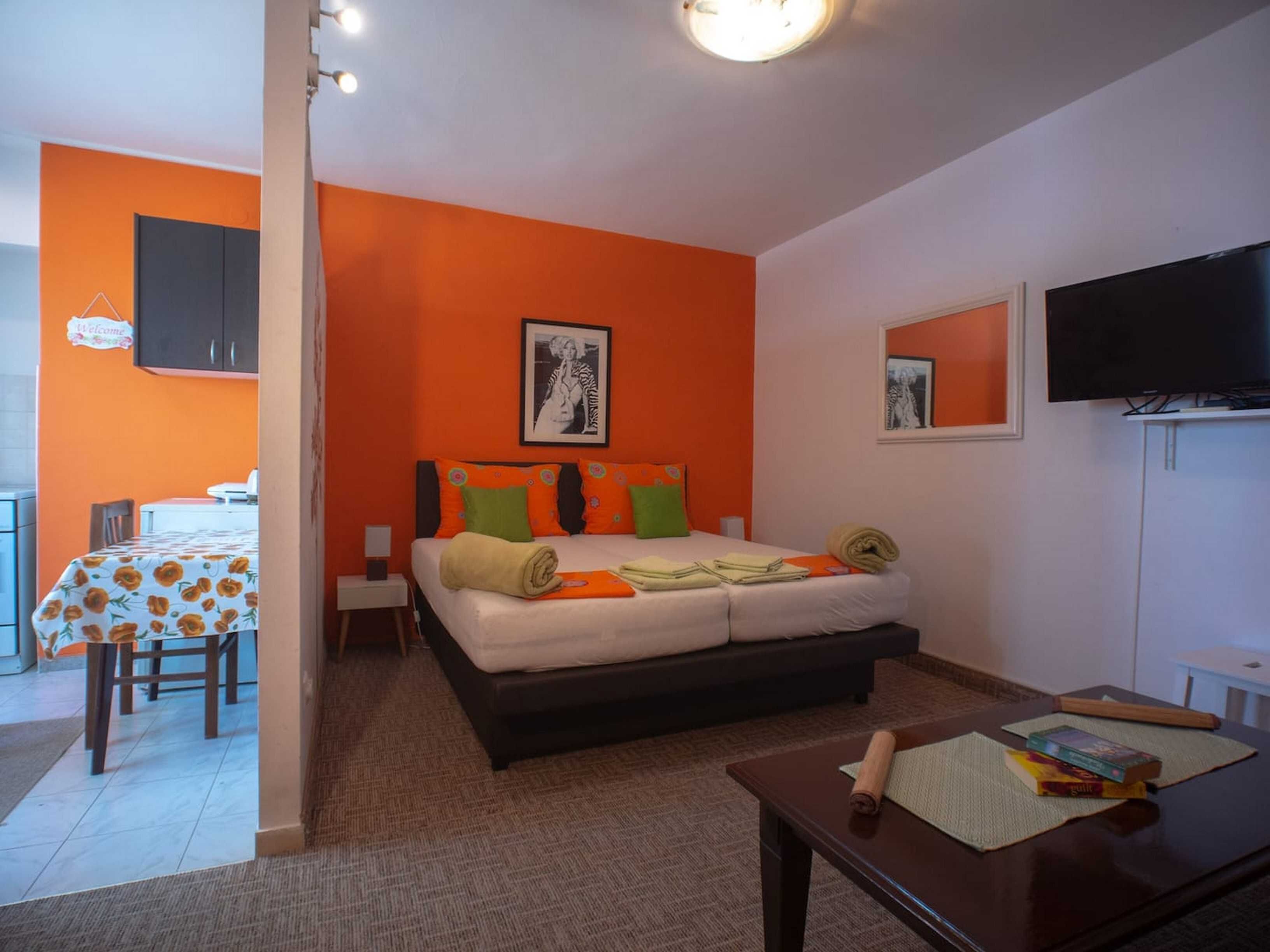 Une chambre avec un mur d’accent orange, un lit avec de nombreux coussins, une table de chevet, une télévision et une petite table avec des livres et un tapis.