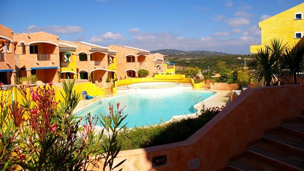 Une cour d’hôtel avec une piscine, des bâtiments aux façades en terre cuite et une vue sur les collines.