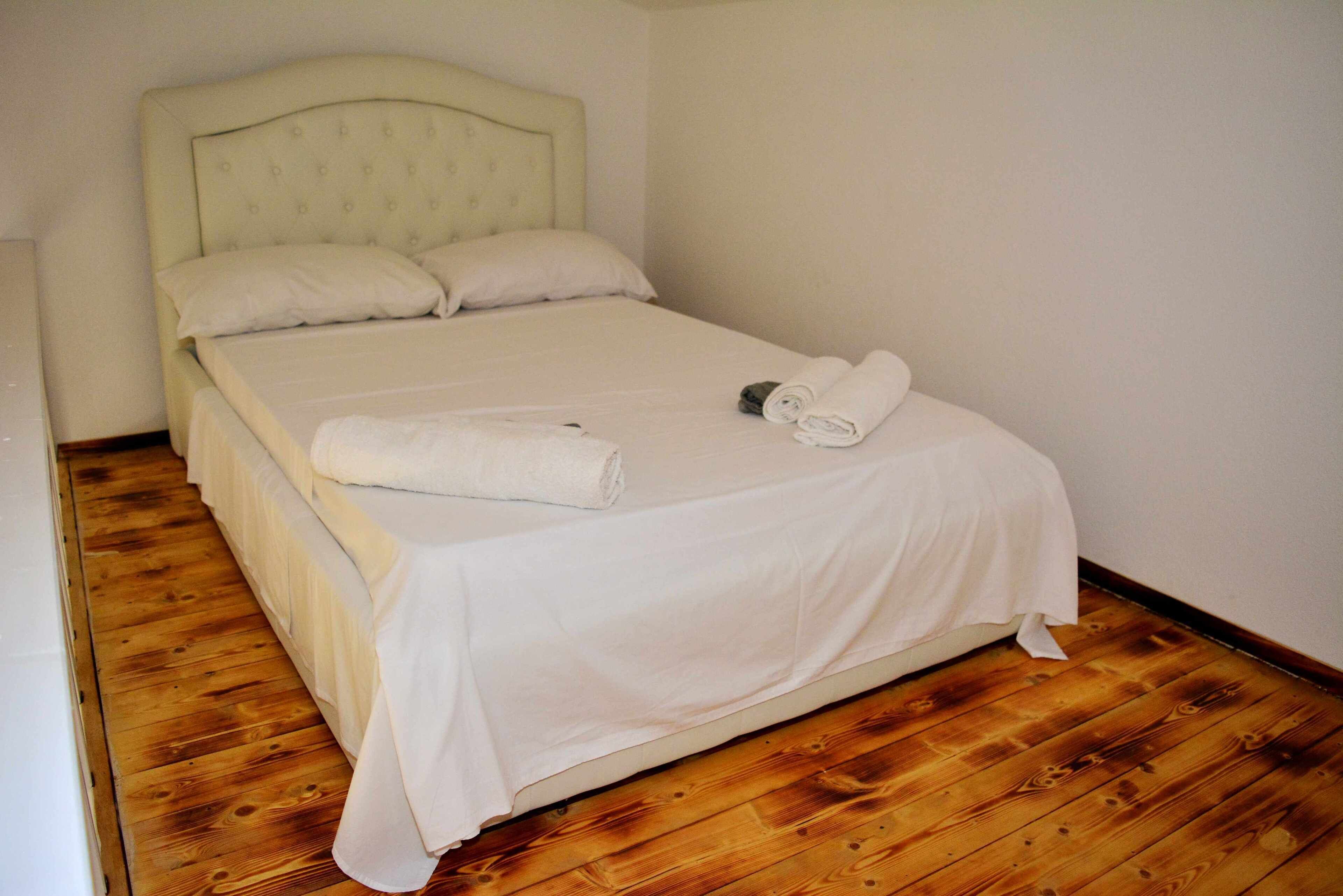 Een net opgemaakt bed met witte lakens en twee opgerolde handdoeken aan de zijkant, in een kamer met houten vloer en een eenvoudig hoofdbord.