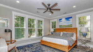 Ein Schlafzimmer mit einem großen Bett, einem Deckenventilator, mehreren Fenstern mit Jalousien und Blick auf Grünfläche.
