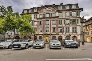 Ein mehrgeschossiges Gebäude im klassischen Stil, mit einem zentralen Eingang und einem dekorativen Bogen. Vor dem Gebäude befinden sich mehrere parkende Autos.