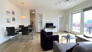 A modern living room with a dining area, a TV, and a large window offering a view of the outside. - BOW-1.25, 6 pers., 70-80sqm - Residence Bollwark 1.2 (Kappeln)