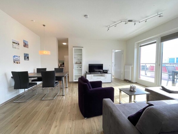 A modern living room with a dining area, a TV, and a large window offering a view of the outside. - BOW-1.25, sleeps 6, 70–80 m² - Bollwark 1.2 Residence (Kappeln)