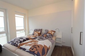 A bedroom with a bed, a nightstand, a window, and a wall-mounted shelf.