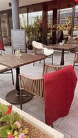 Un área de café al aire libre con sillas y mesas de mimbre, un cartel de menú y un arreglo floral.