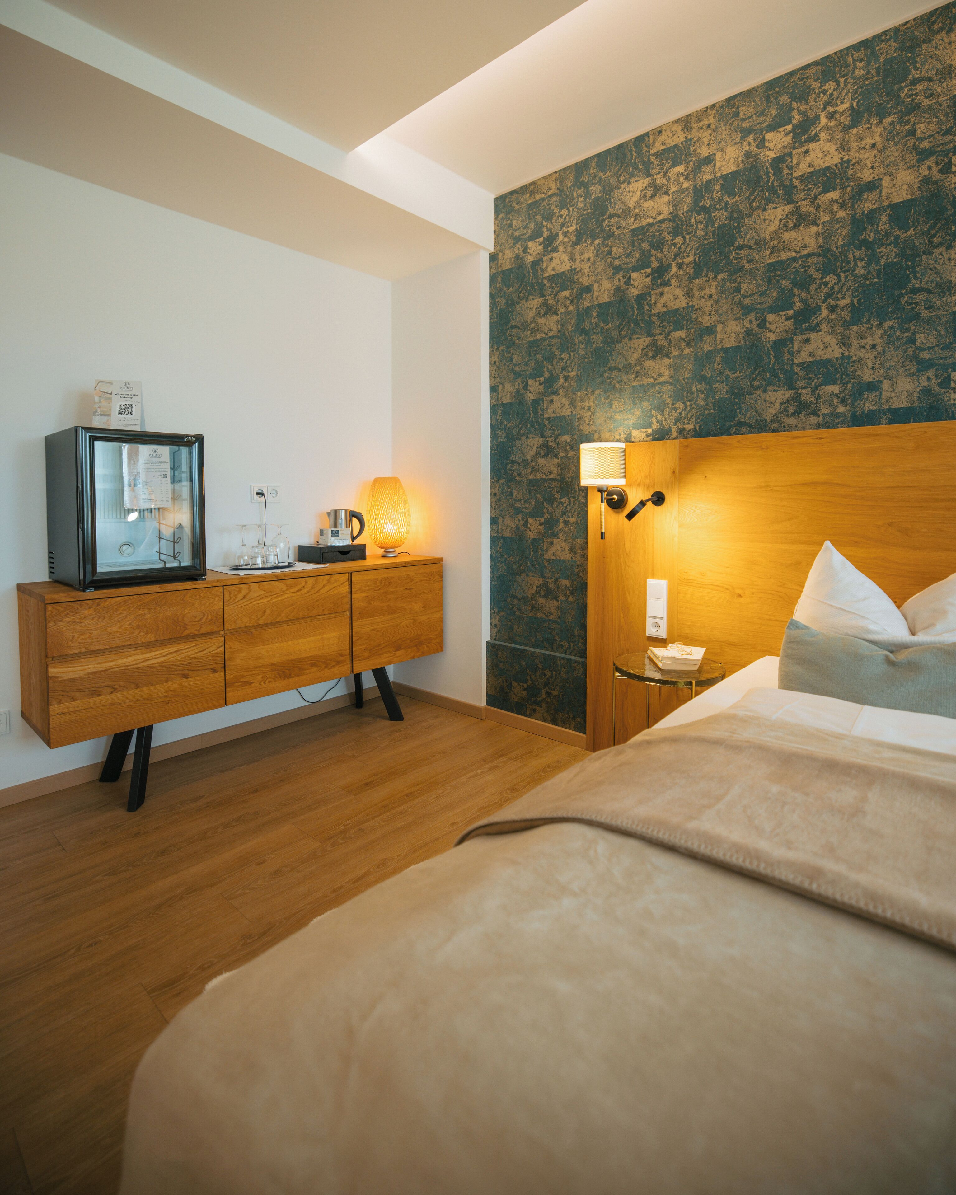 Une chambre d’hôtel avec une commode en bois, un lit recouvert d’une couverture beige, une lampe fixée au mur et un mur à texture.
