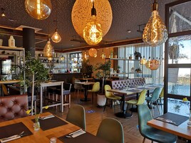 Interior de restaurante con varias mesas, sillas, lámparas colgantes y una barra con estantes llenos de botellas y vasos.