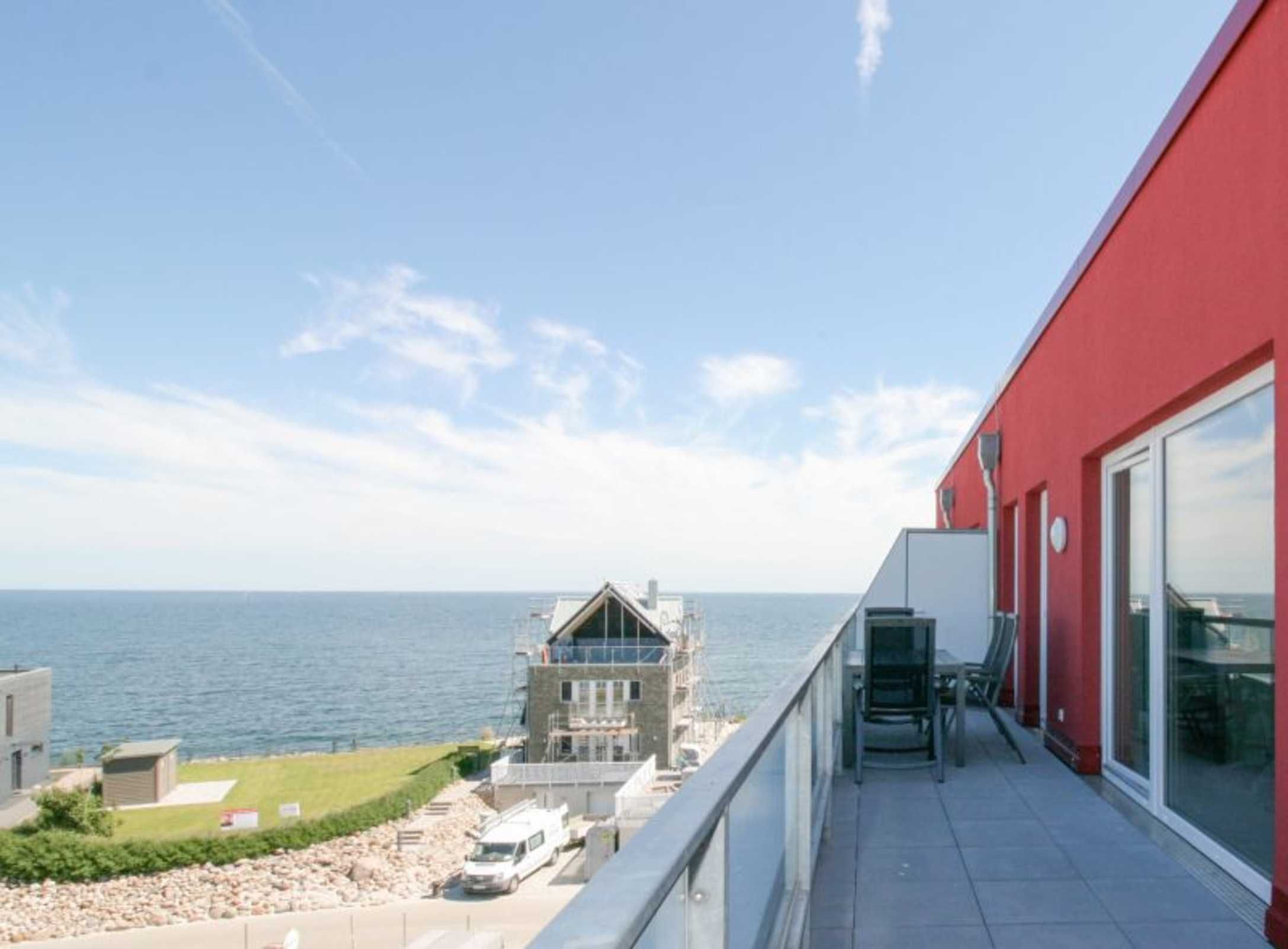 Un edificio moderno con fachada roja, un balcón con barandilla de cristal y vista al mar.
