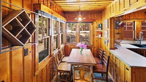 Interior d'una cabana de fusta amb taula de menjador, cadires, zona de cuina i grans finestres.