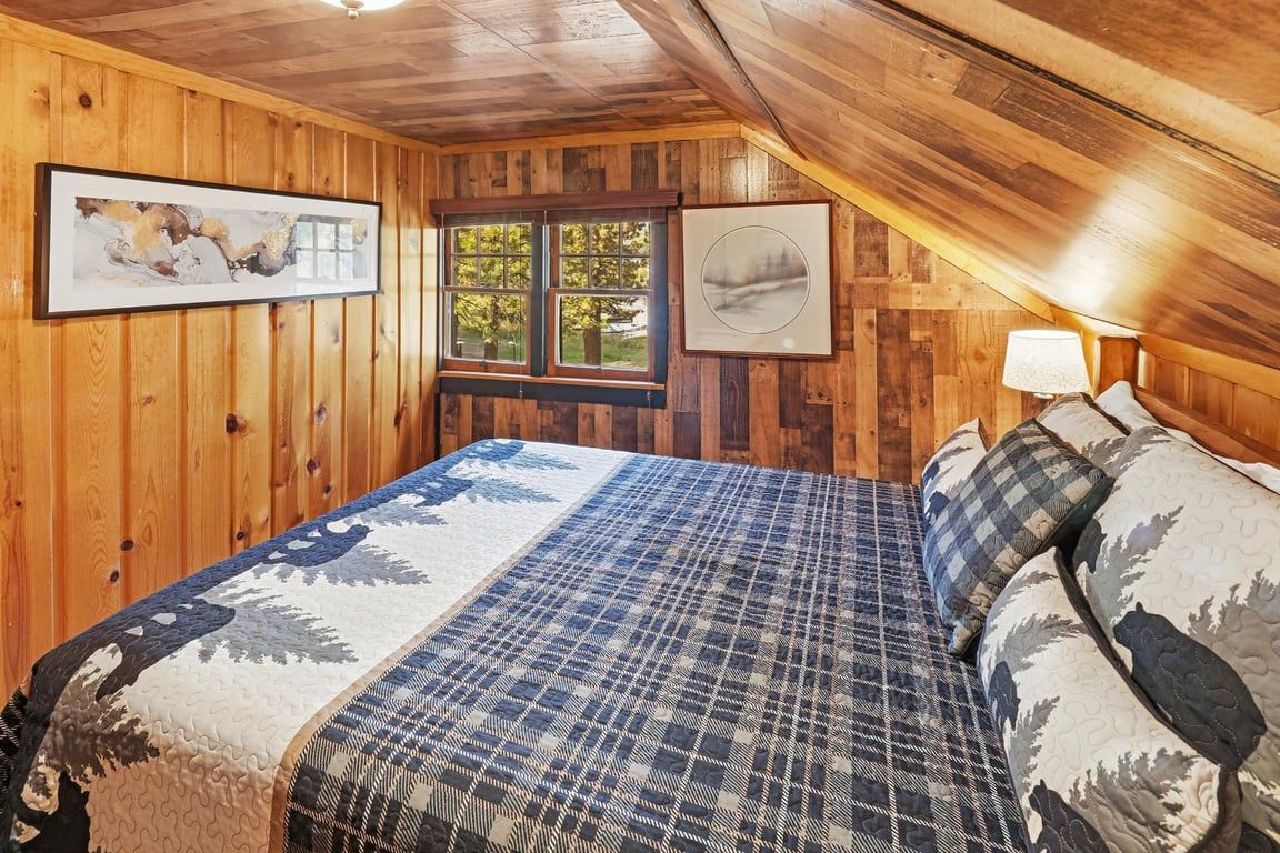 A bedroom with wooden walls, a bed with patterned bedding, a window, and two framed pictures.