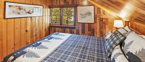 A bedroom with wooden walls, a bed with patterned bedding, a window, and two framed pictures.