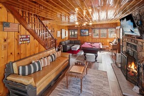 Een gezellig interieur van een chalet met een houten trap, een biljarttafel, een open haard en een comfortabele zithoek.