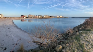 Una zona costera con una playa de arena, una masa de agua tranquila y una hilera de edificios al fondo.