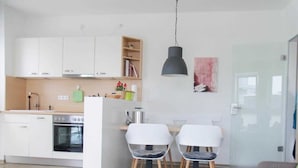 Una cocina moderna con armarios blancos, horno empotrado y una zona de comedor con dos sillas y una mesa blancos.