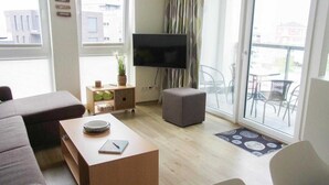 Una sala de estar moderna con un sofá, un reposapiés, una mesa de centro y un televisor montado en la pared. Hay una puerta corrediza de vidrio que da a un área exterior con sillas.