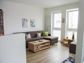 Ein modernes Wohnzimmer mit einer Couch, einem Couchtisch und einem kleinen Beistelltisch. Zwei Fenster sorgen für natürliches Licht, und eine Heizung ist an der Wand zu sehen.