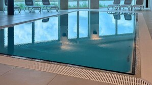 An indoor swimming pool with clear views of a river and distant landscape, surrounded by lounge chairs and large windows.