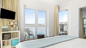 A bedroom with a large bed, a TV mounted on the wall, a chair, a small table, and a view of the city through large windows.