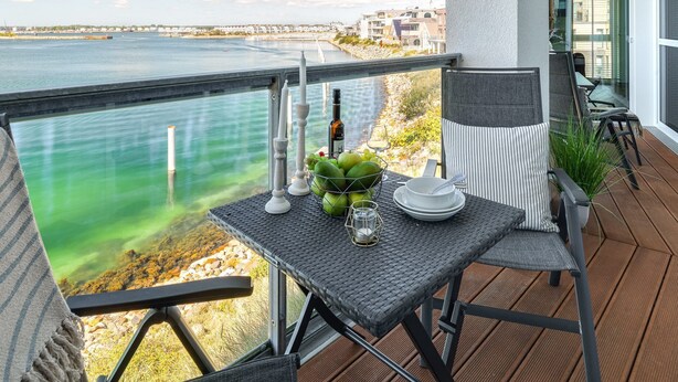 A balcony with a table set for two, overlooking a body of water and a coastal town.