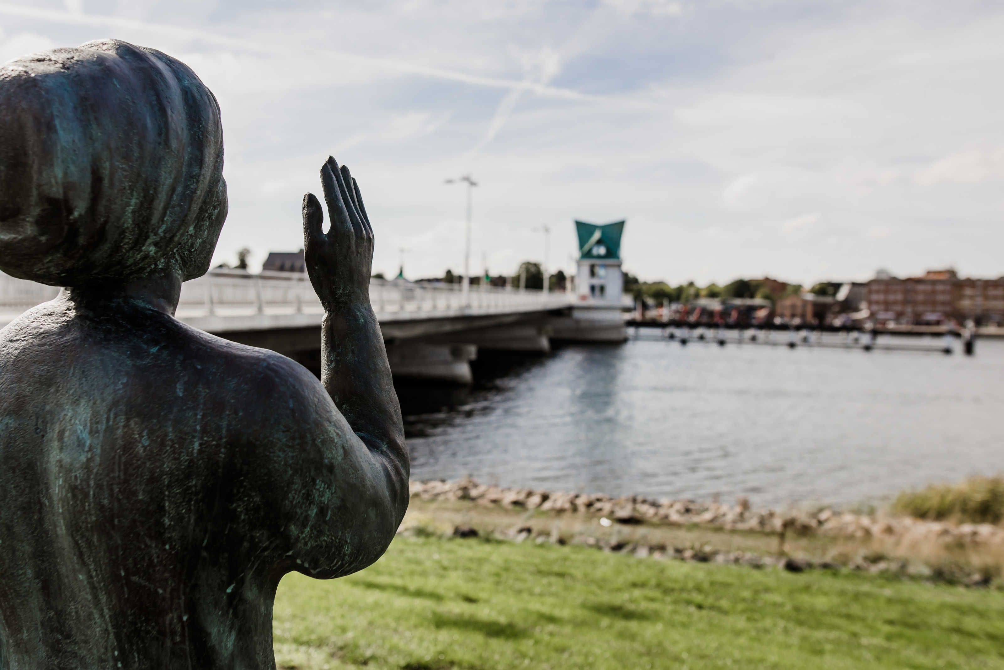 Une statue représentant une personne les bras levés, au bord d’une rivière, avec un pont et des bâtiments en arrière-plan.
