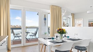 A dining area with a white table set for two, a balcony with a view of the water, and a cityscape in the background.